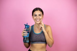 Woman in sports clothes holding a water bottle and smiling while pointing to her jaw